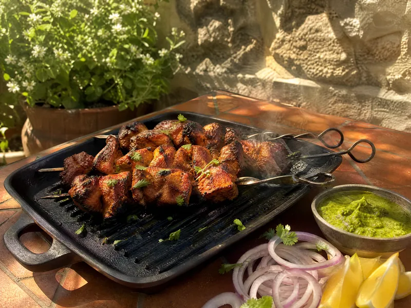 Charred chicken tikka on skewers with onion rings, lemon and green mint chutney on a terracotta table.