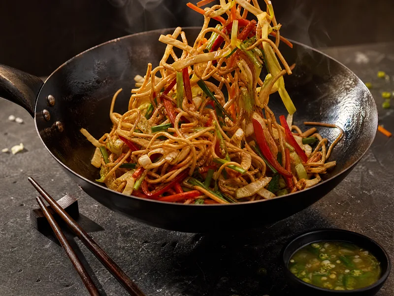 A black wok of vegetable Hakka noodles with shredded cabbage, carrot, peppers and spring onion, with chopsticks.