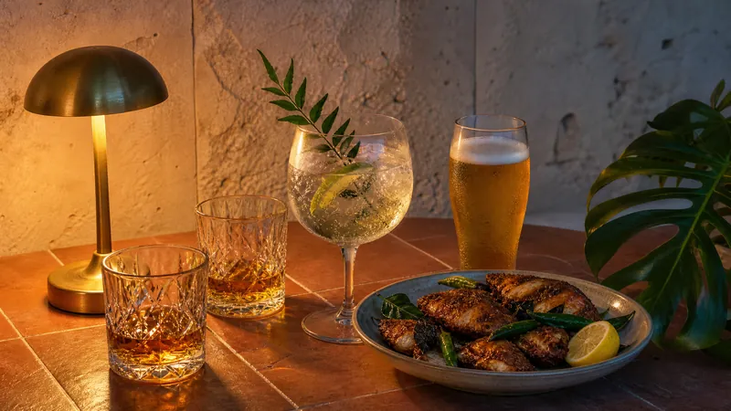 The same table at dusk with whisky, a gin and tonic, a lager and spiced fried fish under a brass lamp.