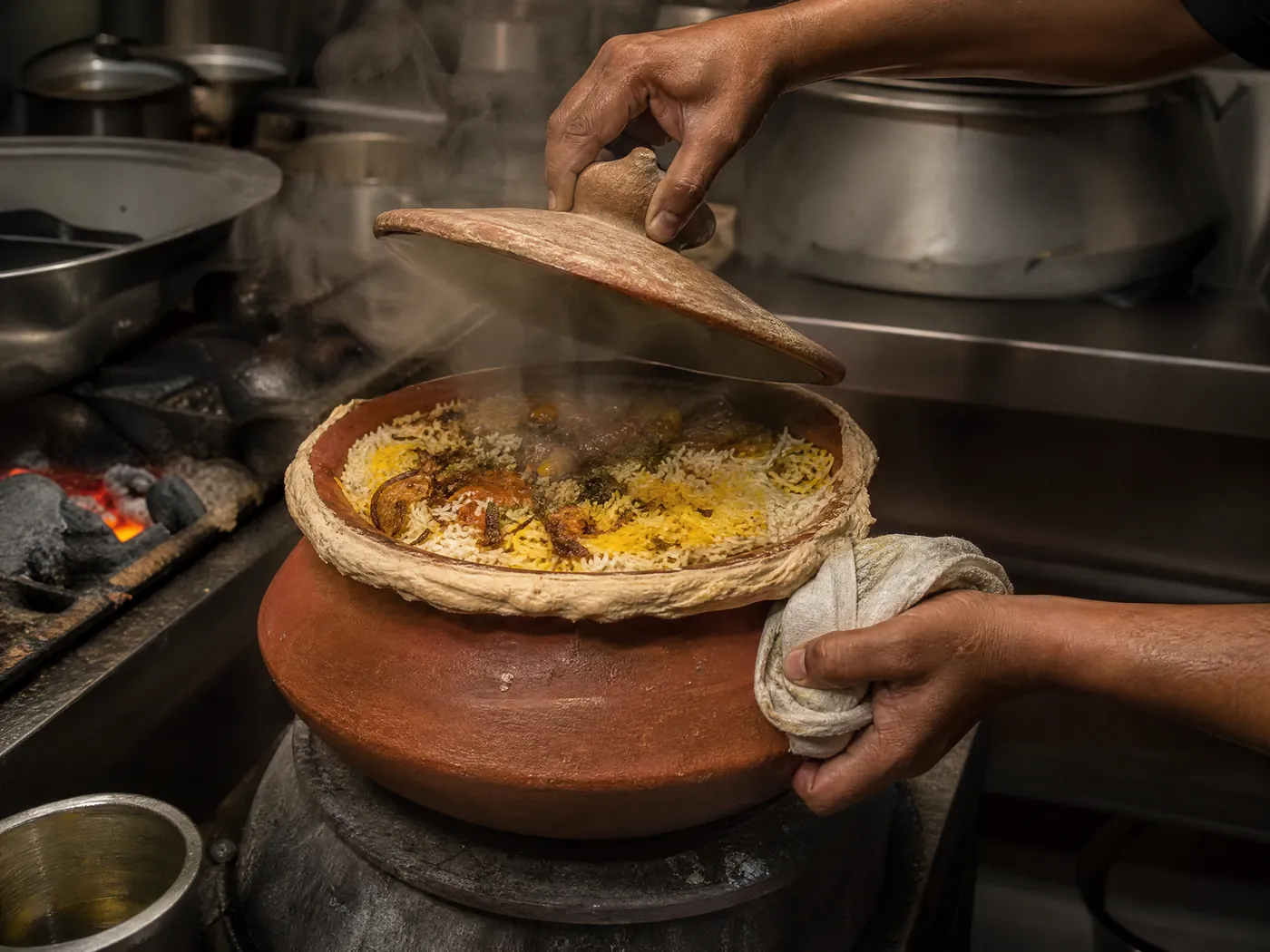 A cook lifts the lid from a dough-sealed clay pot of biryani.